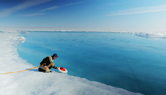 A man kneels at the edge of an ice sheet and releases a small boat into a large body of blue water