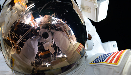 Gloved hands and a camera are reflected in the gold visor of astronaut Kate Rubins during her spacewalk