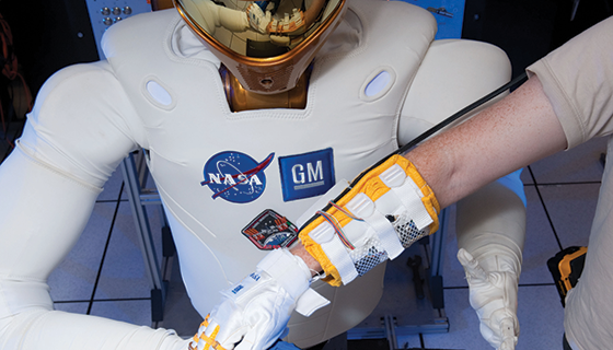 Two people wearing robo-gloves each place one of their hands onto Robonaut2's hand