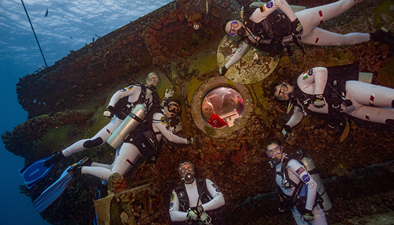 Two crew members peer out the window of an underwater habitat as six crew members in scuba gear pose outside