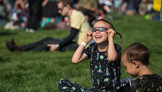 A boy wearing a rocket-patterned outfit smiles while looking through eclipse glasses