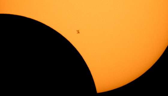 The space station looks like a small speck in front of a partially eclipsed sun