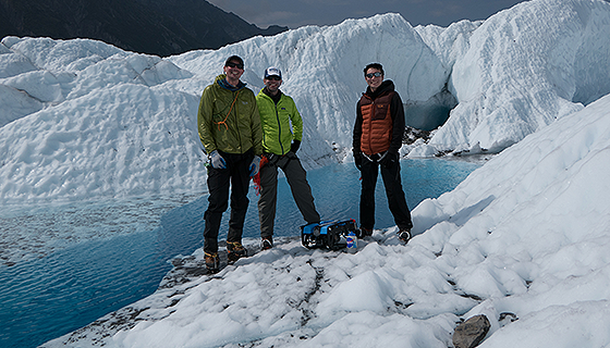 Three men stand near a small robot on the edge of a pool of water surrounded by ice