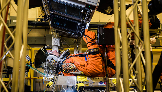 A person in a spacesuit lies in a simulator with controls overhead