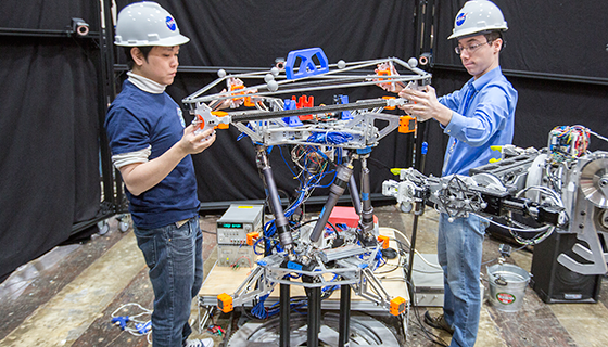 Two engineers wearing hard hats hold pieces of metal being assembled by a robotic arm