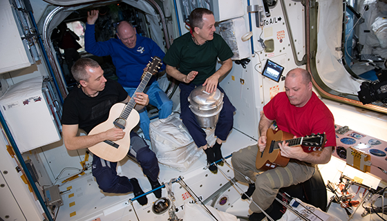 Two astronauts play guitars and one plays a drum while floating inside the space station