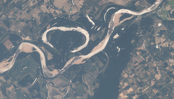 A view from space of the brown waters of the Mississippi River winding through plowed and green fields with forests