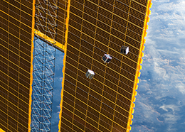 Three small cube-shaped satellites, in the center of the picture, float past a space station solar array