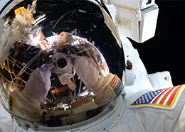 Gloved hands and a camera are reflected in the gold visor of astronaut Kate Rubins during her spacewalk