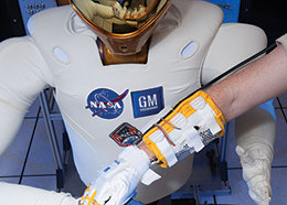 Two people wearing robo-gloves each place one of their hands onto Robonaut2's hand