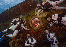 Two crew members peer out the window of an underwater habitat as six crew members in scuba gear pose outside