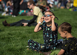 A boy wearing a rocket-patterned outfit smiles while looking through eclipse glasses