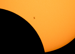 The space station looks like a small speck in front of a partially eclipsed sun
