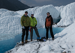 Three men stand near a small robot on the edge of a pool of water surrounded by ice