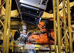 A person in a spacesuit lies in a simulator with controls overhead