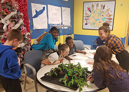 Smiling students surround a table as they record data next to a tray of plants