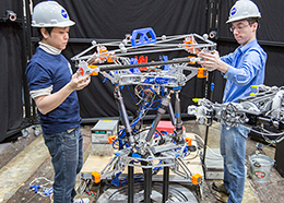 Two engineers wearing hard hats hold pieces of metal being assembled by a robotic arm