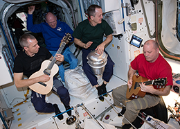 Two astronauts play guitars and one plays a drum while floating inside the space station