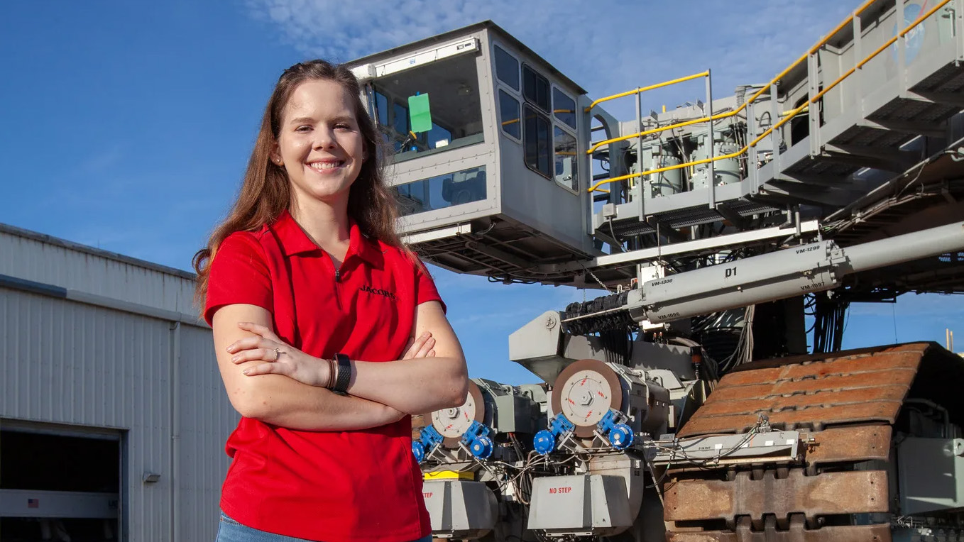 Breanne Stichler smiles while standing in front of the crawler, a giant machine with large tank treads that is used to transport rockets to a launch pad.