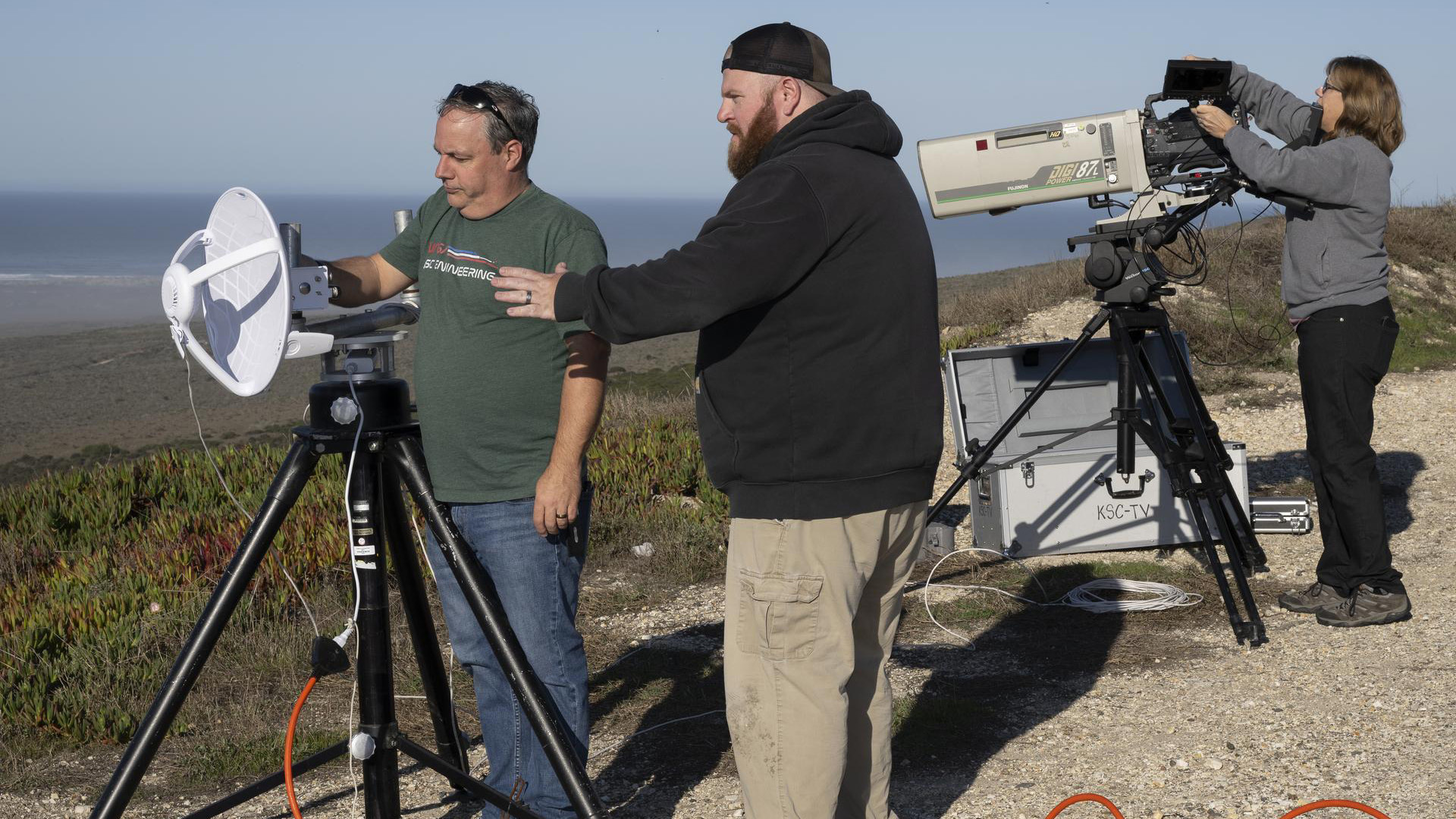 Three videographers set up cameras and microphones on tripods on a hillside in preparation for filming a rocket launch at Vandenberg Space Force Base.