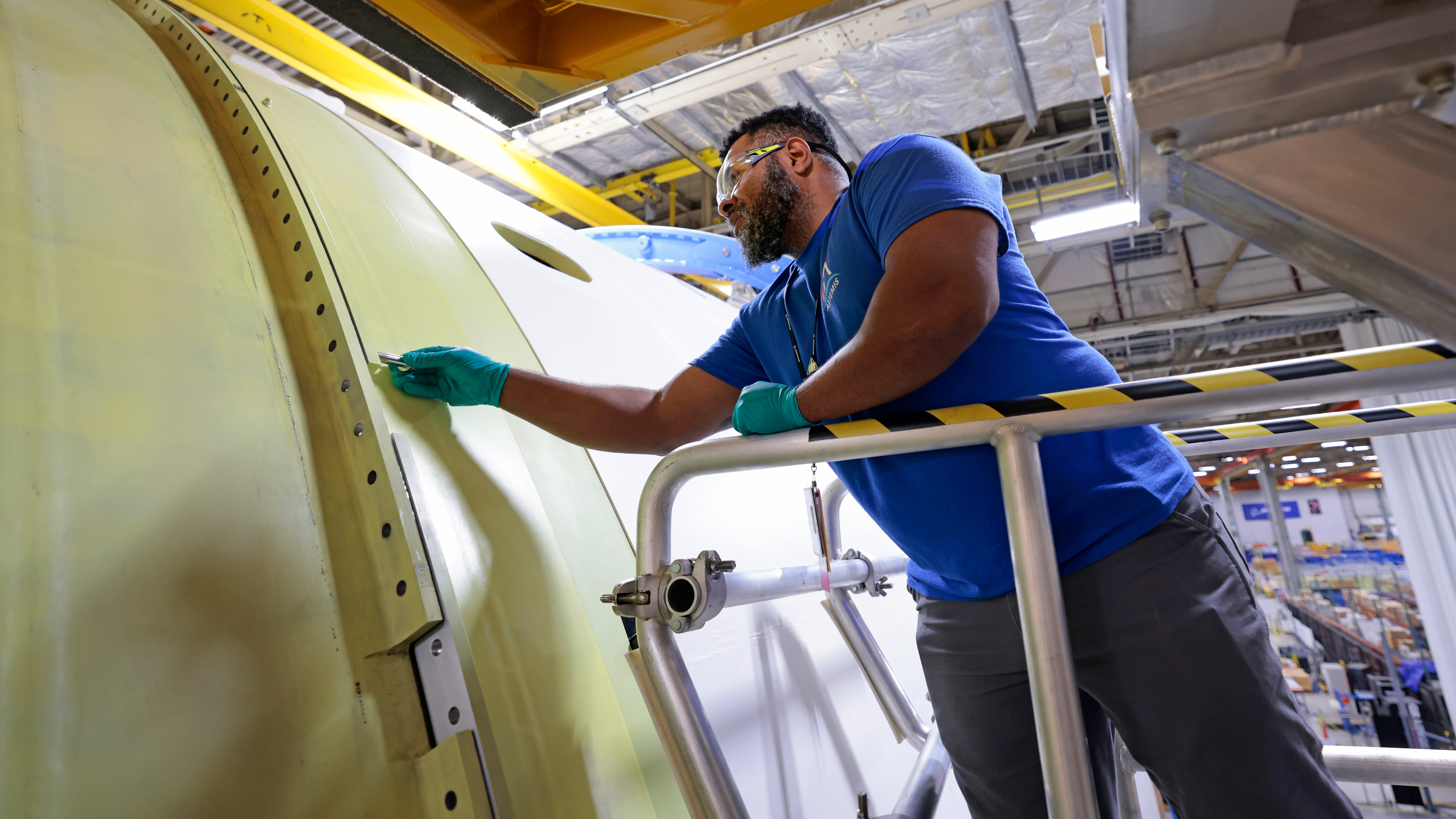 A technician wearing safety glasses and standing on a lift holds out an instrument to check the connection between two large sections of the Space Launch System rocket that will be used for the Artemis II mission.