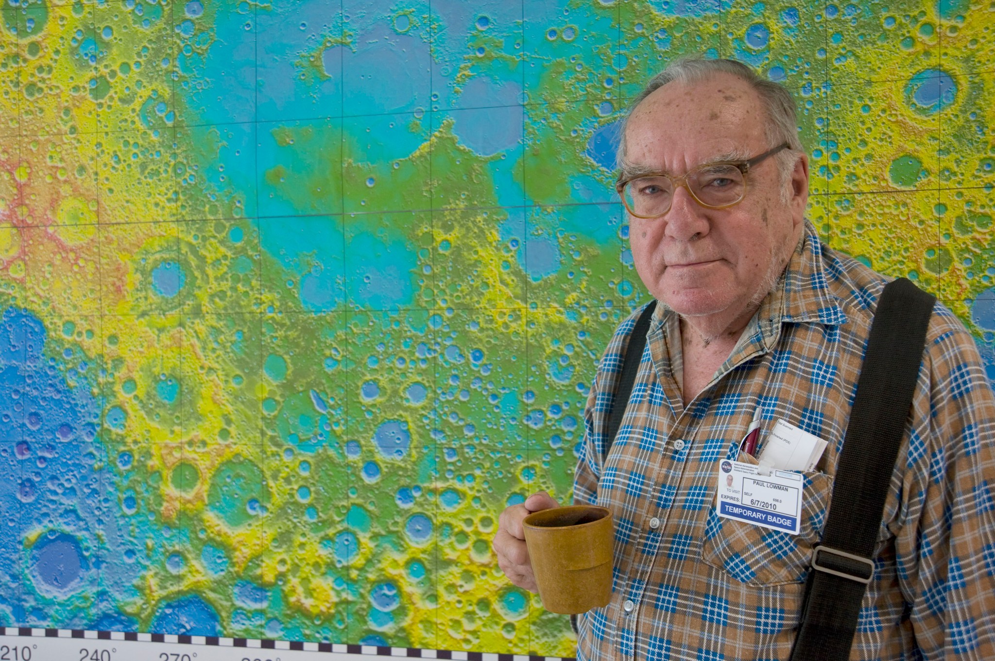 Dr. Paul Lowman wearing suspenders, holding a coffee mug, standing in front of a multicolored lunar map