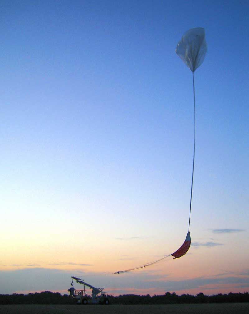 A balloon ascends a twilight sky, with colors from orange near the horizon to deep blue at the top of the frame. Below the balloon is a conical recovery parachute, which is attached to the payload, still attached to a support vehicle at bottom left.