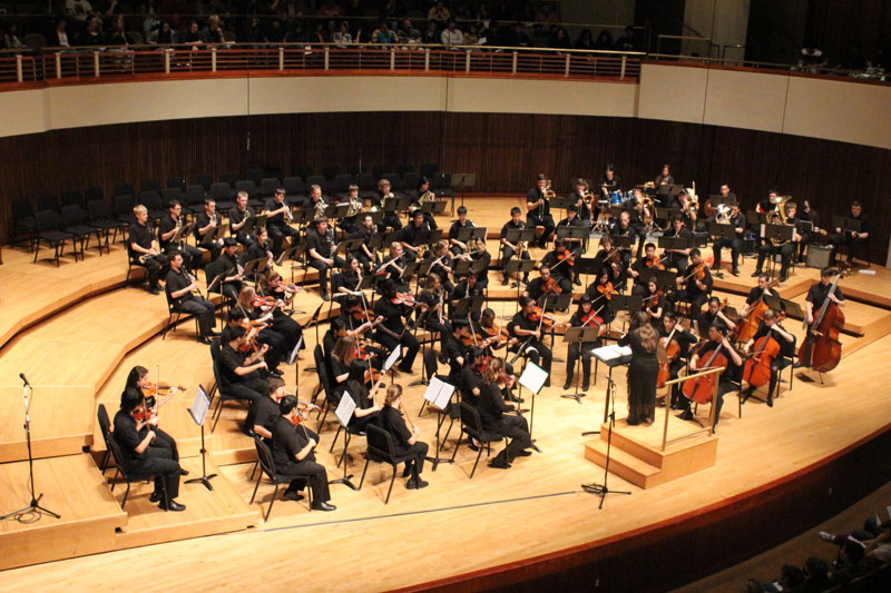 Wide shot of a symphonic orchestra in black uniforms on a tan stage
