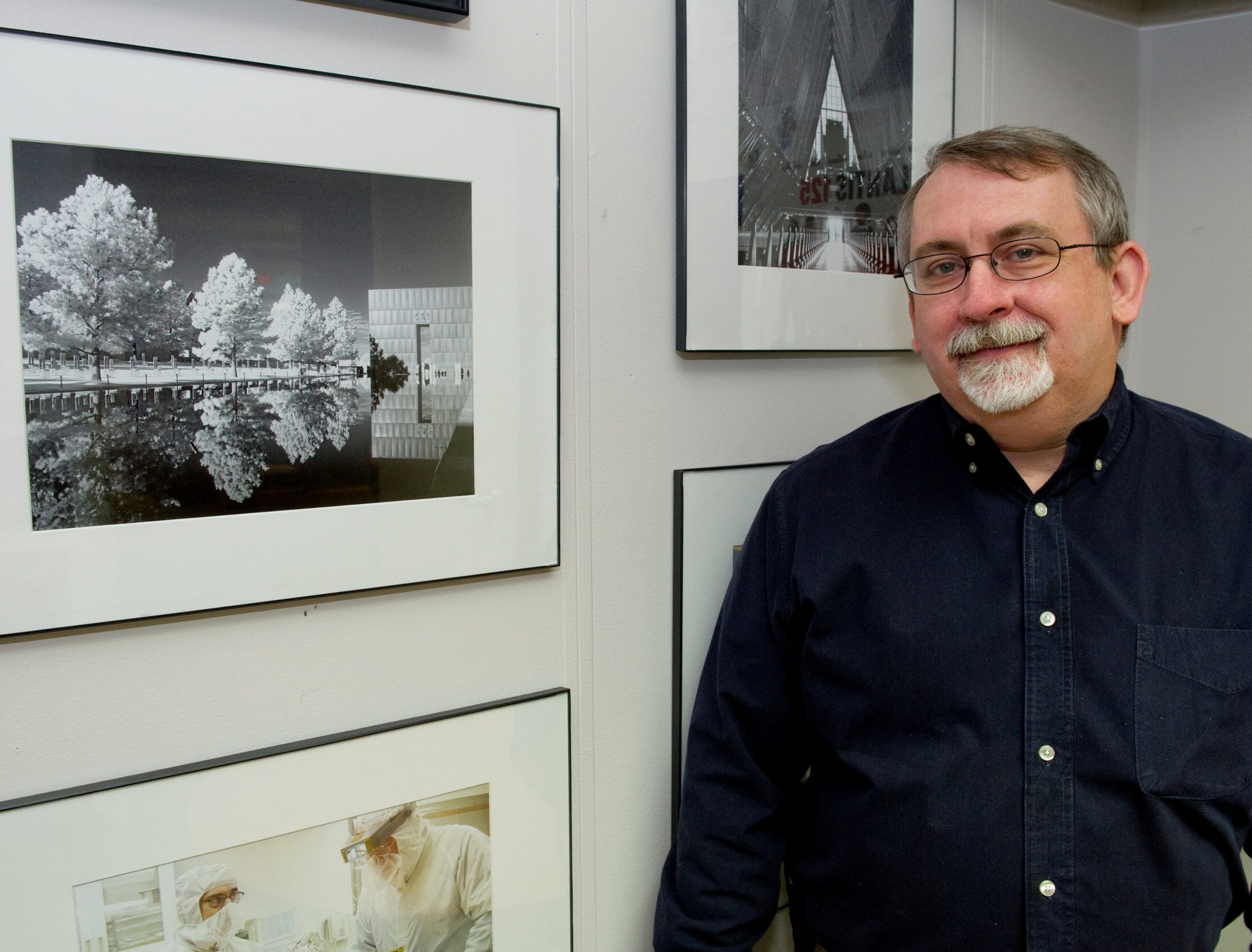 man with glasses and goatee stands by a wall of framed photographs