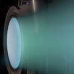 A blue-green glow is emitted from a metal circle as the NEXT engine fires at full power. 