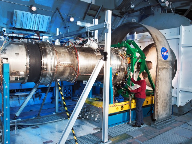 A female engineer stands beside a large engine inside a test facility, checking wiring.