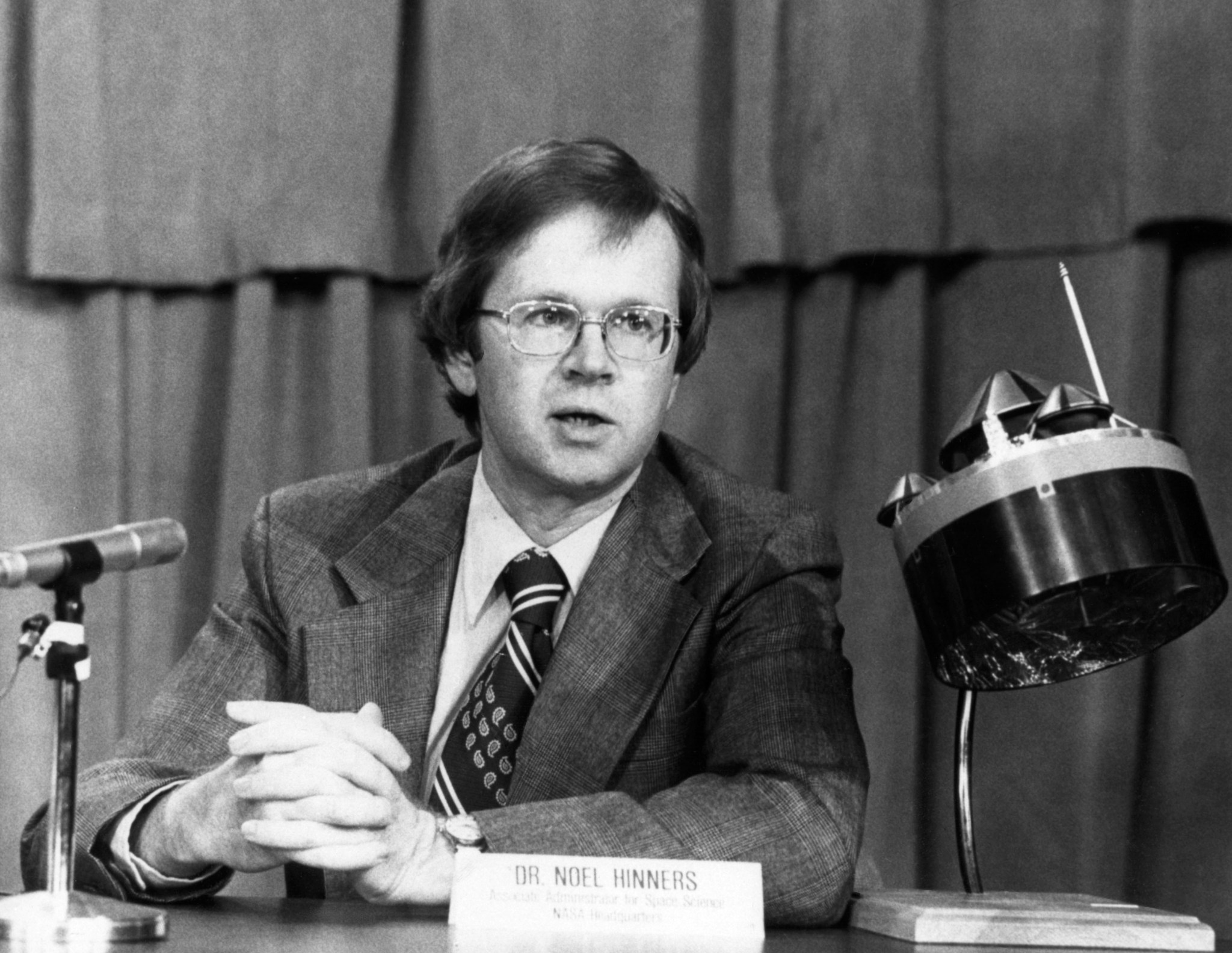 black-and-white photo of Noel Hinners, seated with microphone, satellite model, and name placard