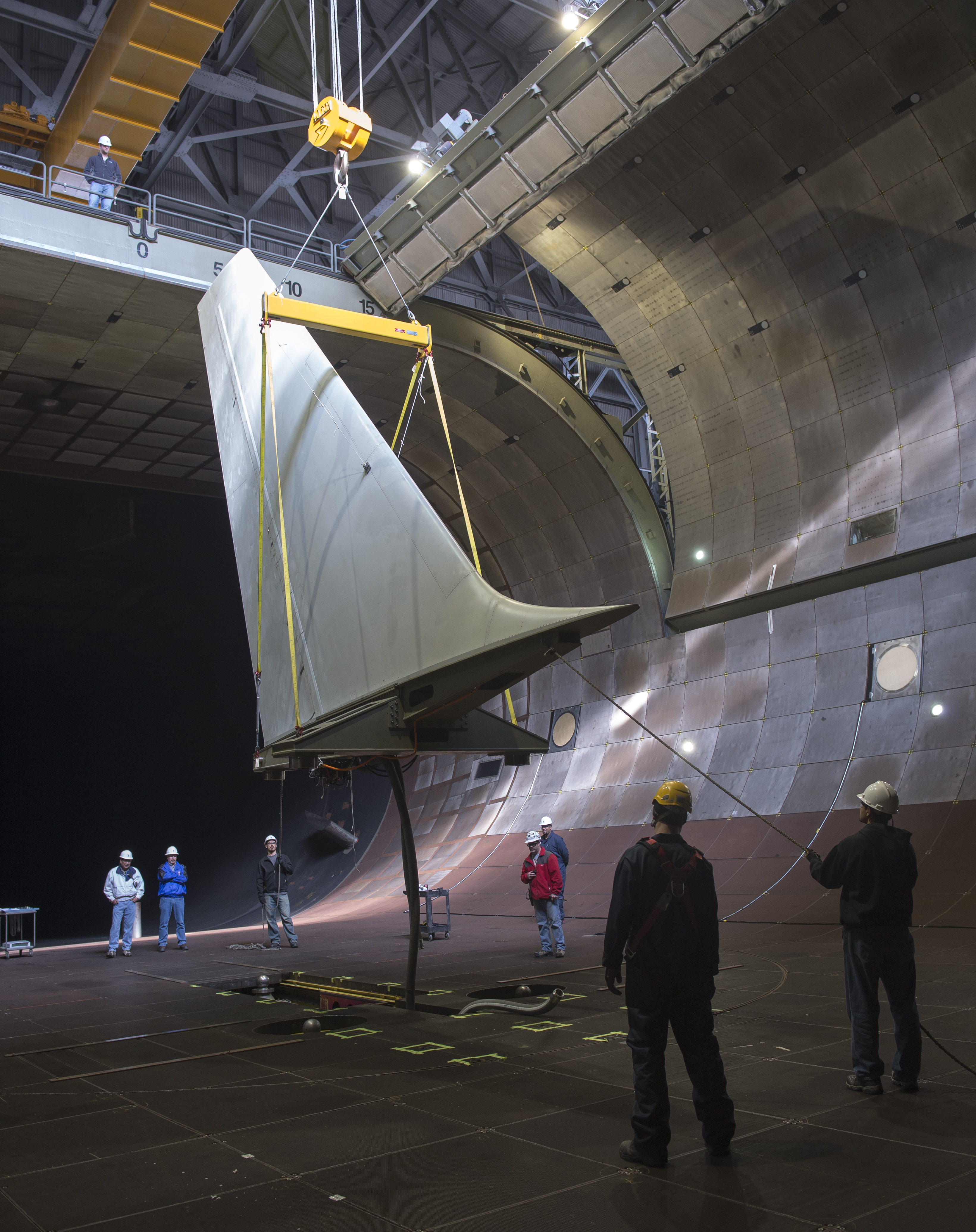 A crane holding a large aircraft tail with workers with hard hats look on.