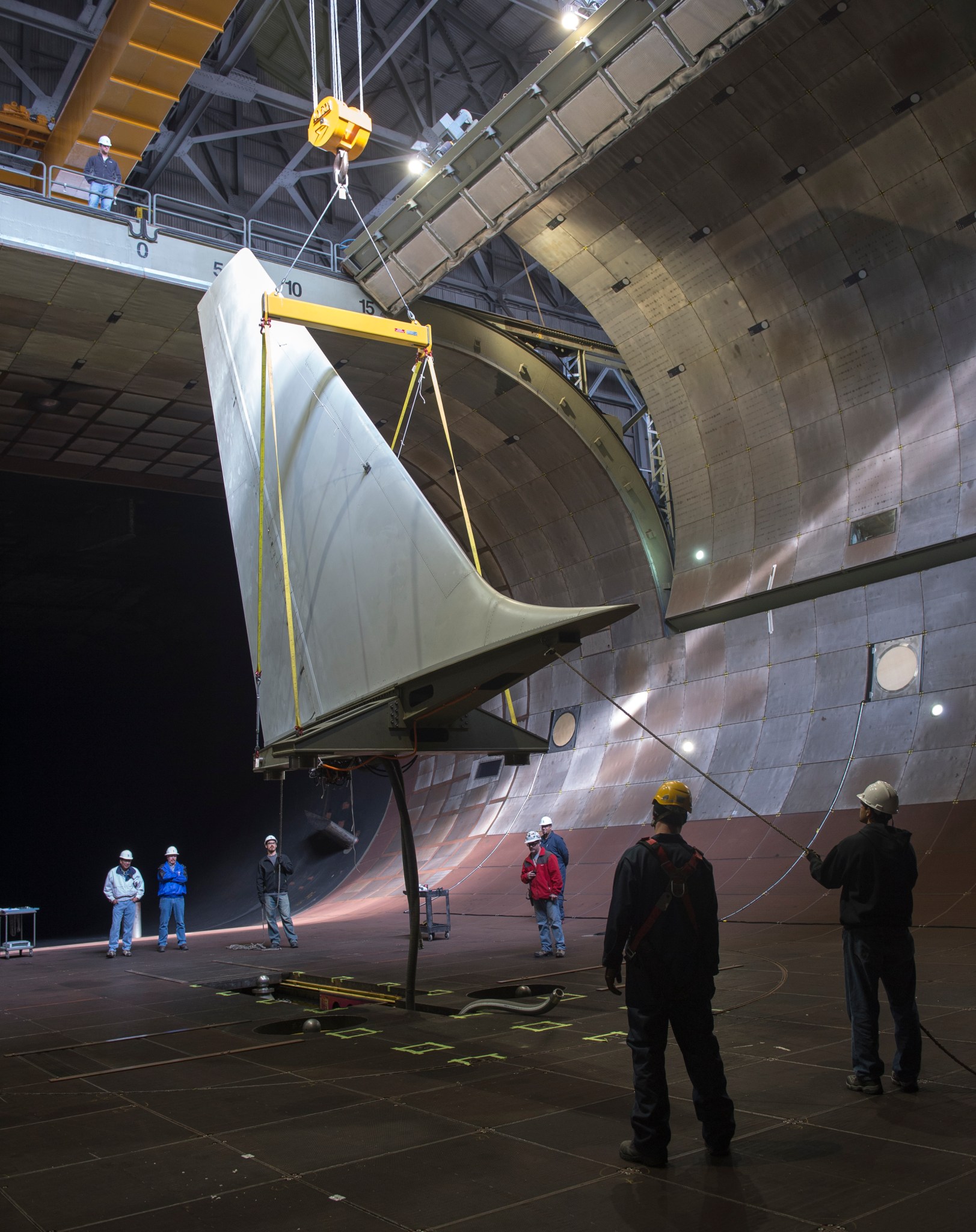 A crane holding a large aircraft tail with workers with hard hats look on.