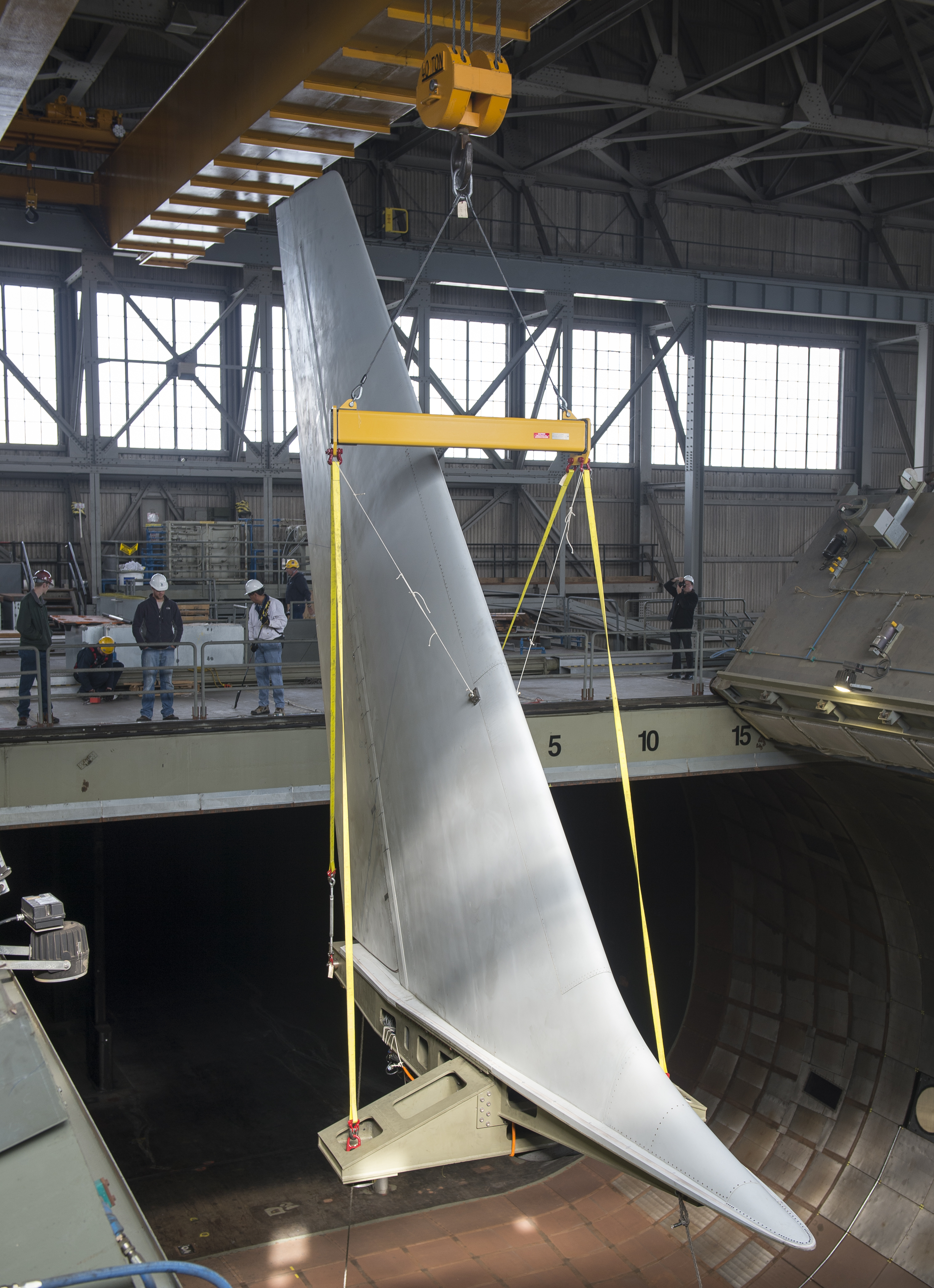 A crane holding a large aircraft tail with workers with hard hats guide the tail.