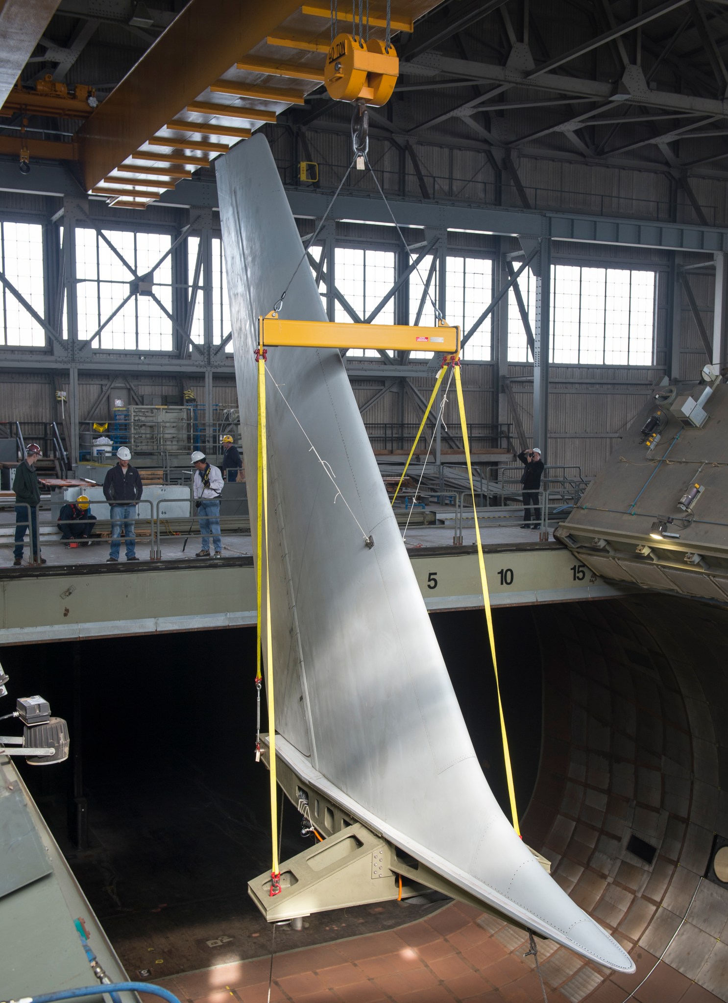 A crane holding a large aircraft tail with workers with hard hats guide the tail.