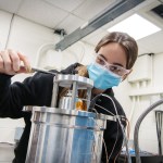 Masked female researcher behind a Stirling converter taking a measurement.