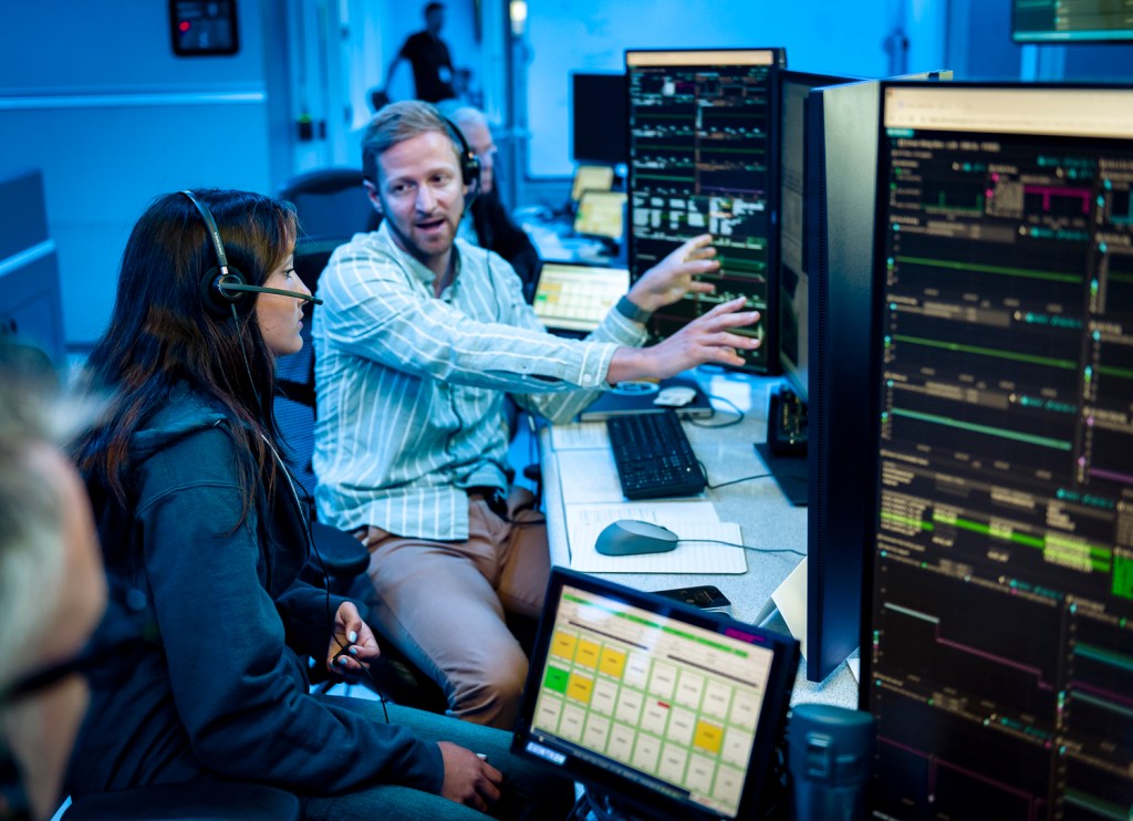 two people sitting infront of screens that look to be control panbels and one person is explaining something to the other person