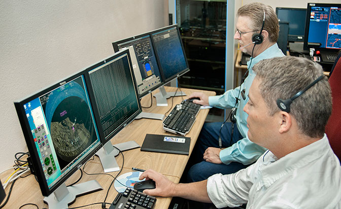 Two men in headsets looking at computer screens.