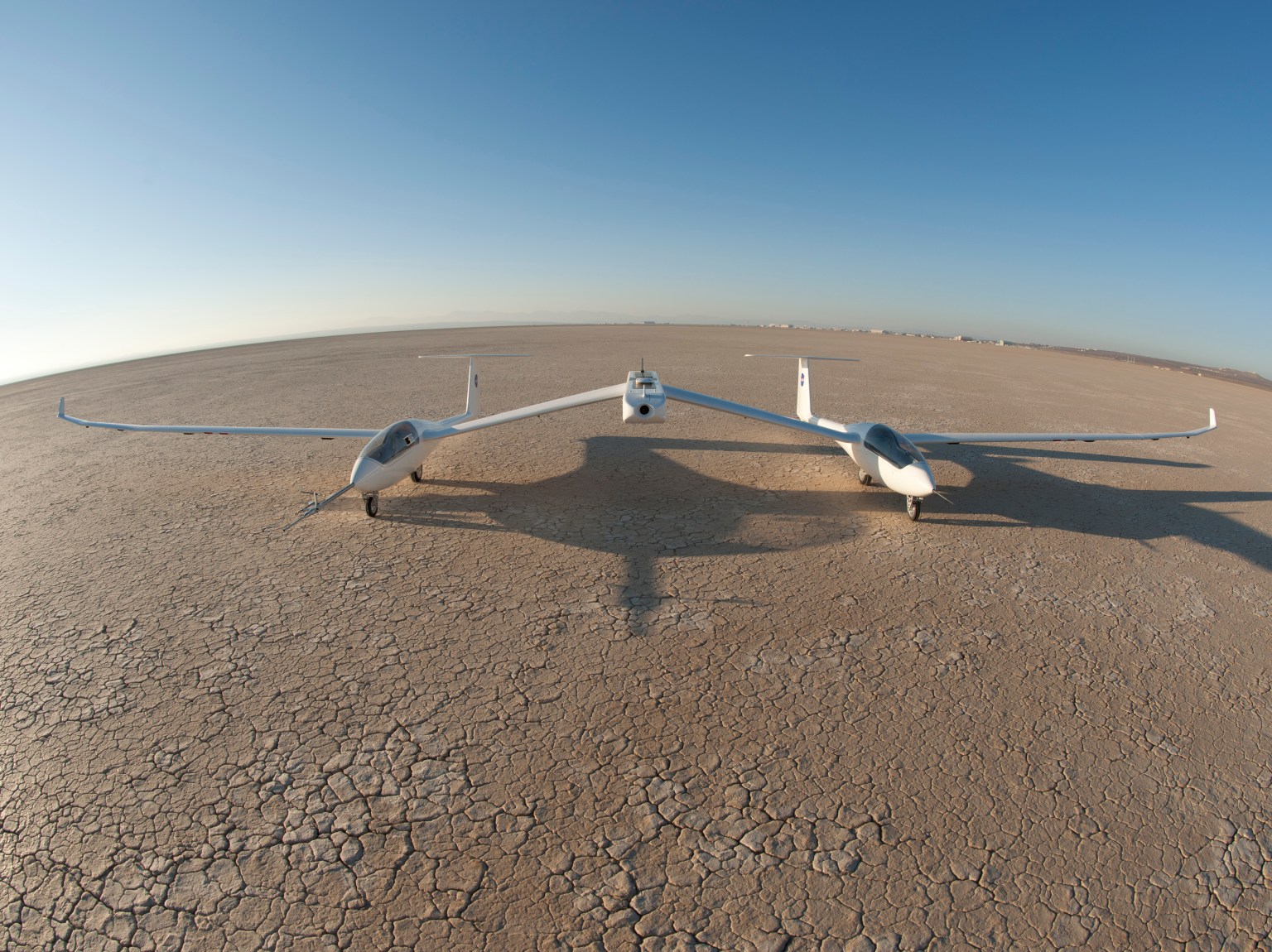 A twin fuselage aircraft sits on a dry lakebed with blue sky in the background.