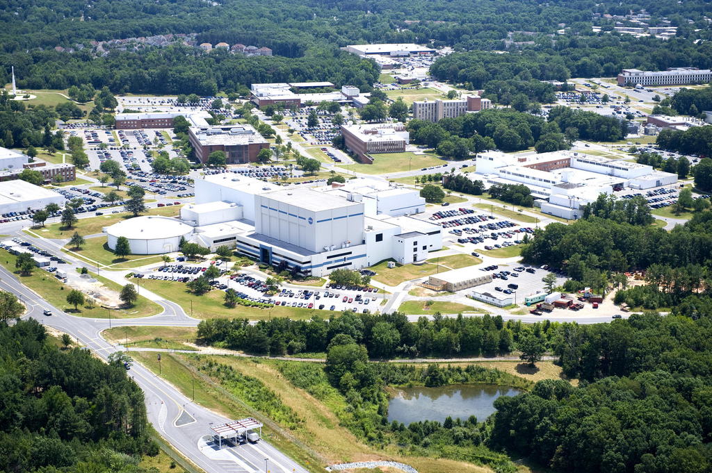 An aerial photograph of the NASA Goddard Visitor Center, with white and brick rectangular buildings peeking out from among dense green trees behind a tall white rocket. The rocket stands in the center of a tan paved circle and is shaped like a pencil with a long, slim tower and a slight bulge at the top. Several smaller red, white, and yellow rockets are displayed on their sides around the paved circle, with another rocket standing in a smaller, white paved circle near the trees.