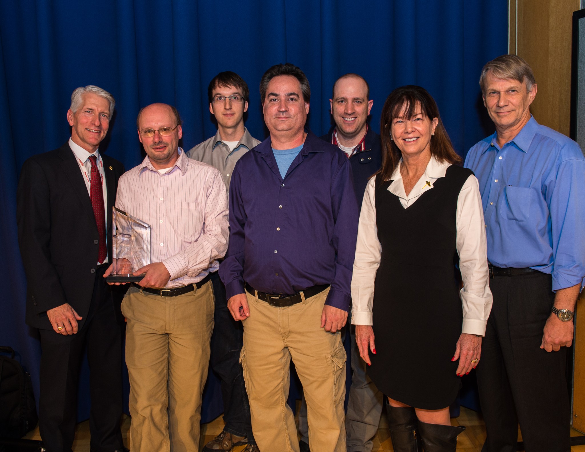 Six men and one woman stand together. The man second to the left wears a pink dress shirt, tan pants and is holding the "Innovator of the year" award
