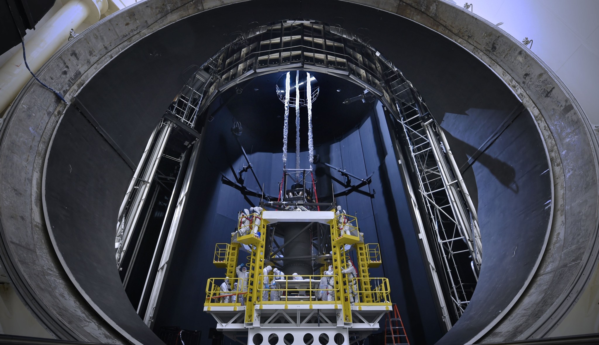 Engineers and technicians, dressed in sterile suits and secured by harnesses to stands for safety, are seen inside Chamber A preparing a lift system that will be used to hold the telescope during testing.