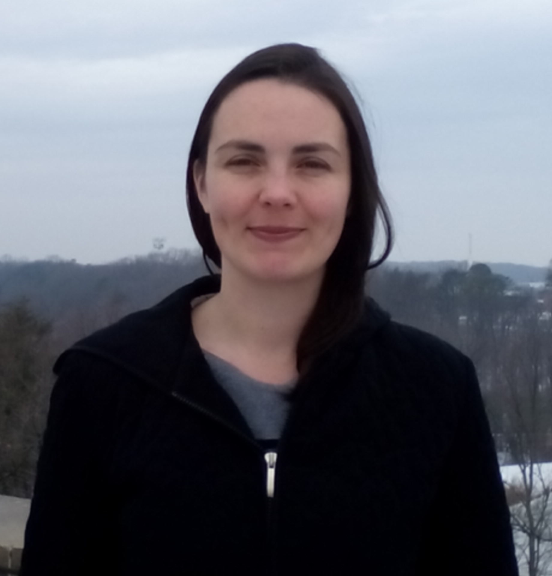 Woman with long brown hair smiles in front of trees and a grey sky. She is wearing a black jacket. 