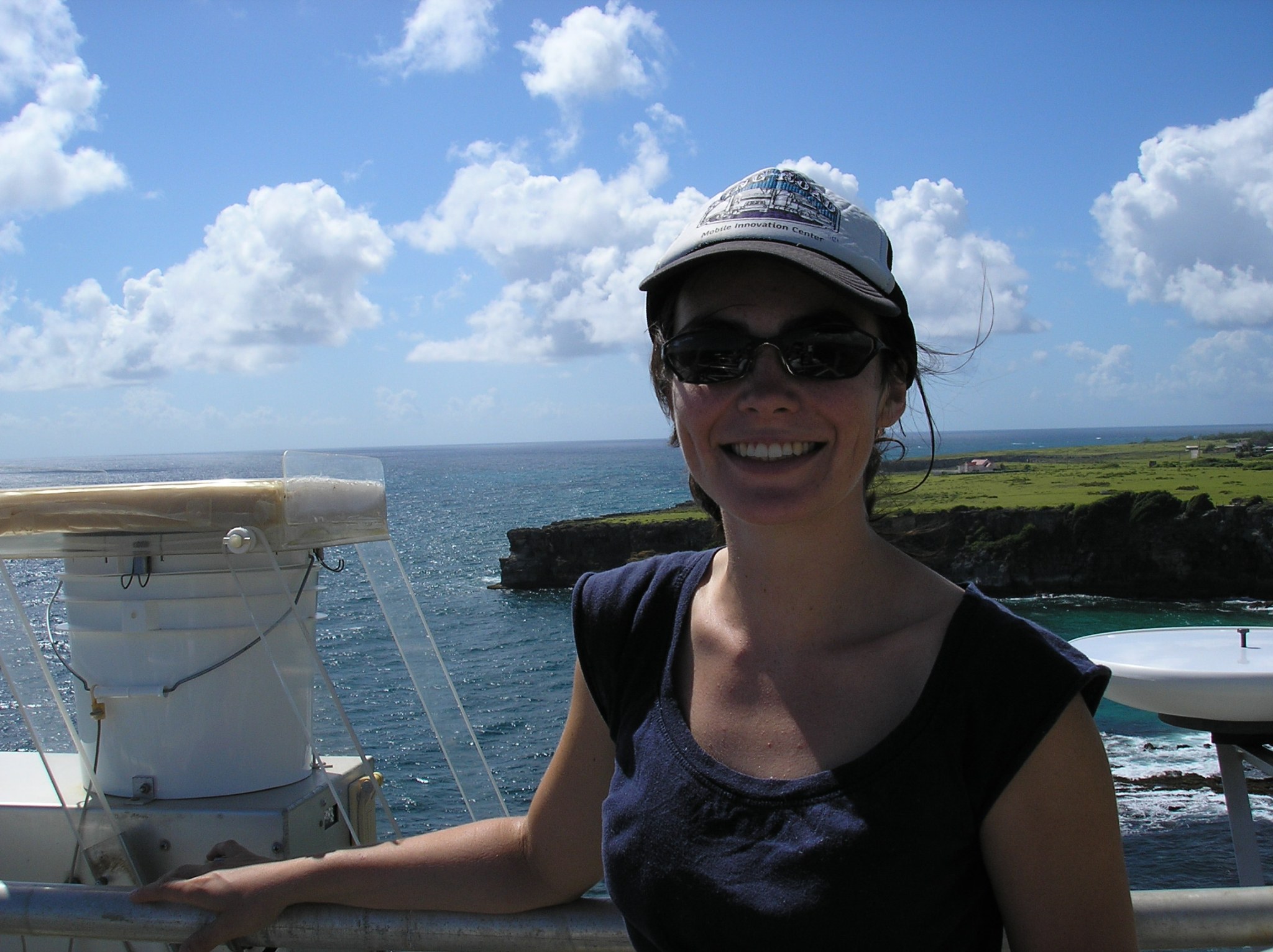Woman with brown hair and fair skin wearing a hat and blue shirt. She is in front of blue water and a green landscape. 