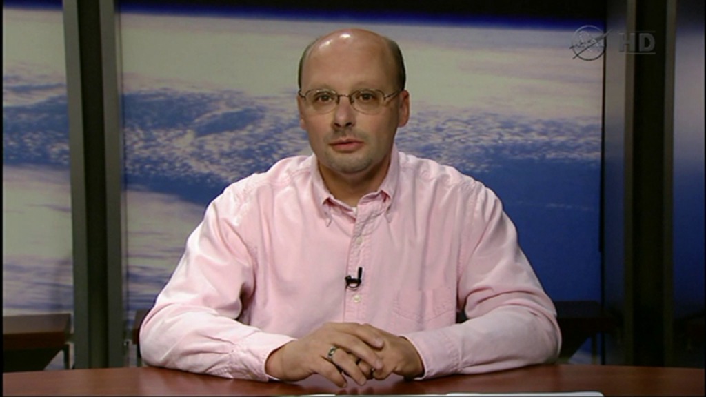 Man with fair skin wears a pink dress shirt, glasses, and a lapel mic.