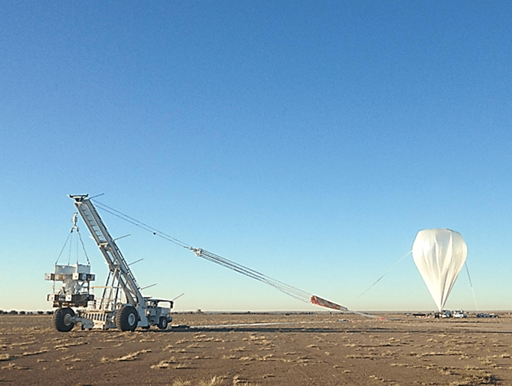 Pale yellow-grey shallow ground takes up only the bottom fifth of the image. In the foreground on the left is the Wallops Arc-Second Pointer (WASP) suspending the Hyper-Spectral Imager for Climate Science (HySICS). The WASP/HySICS is tethered to a large white balloon in the background in the far right of the image. The top half of the image is blue sky.