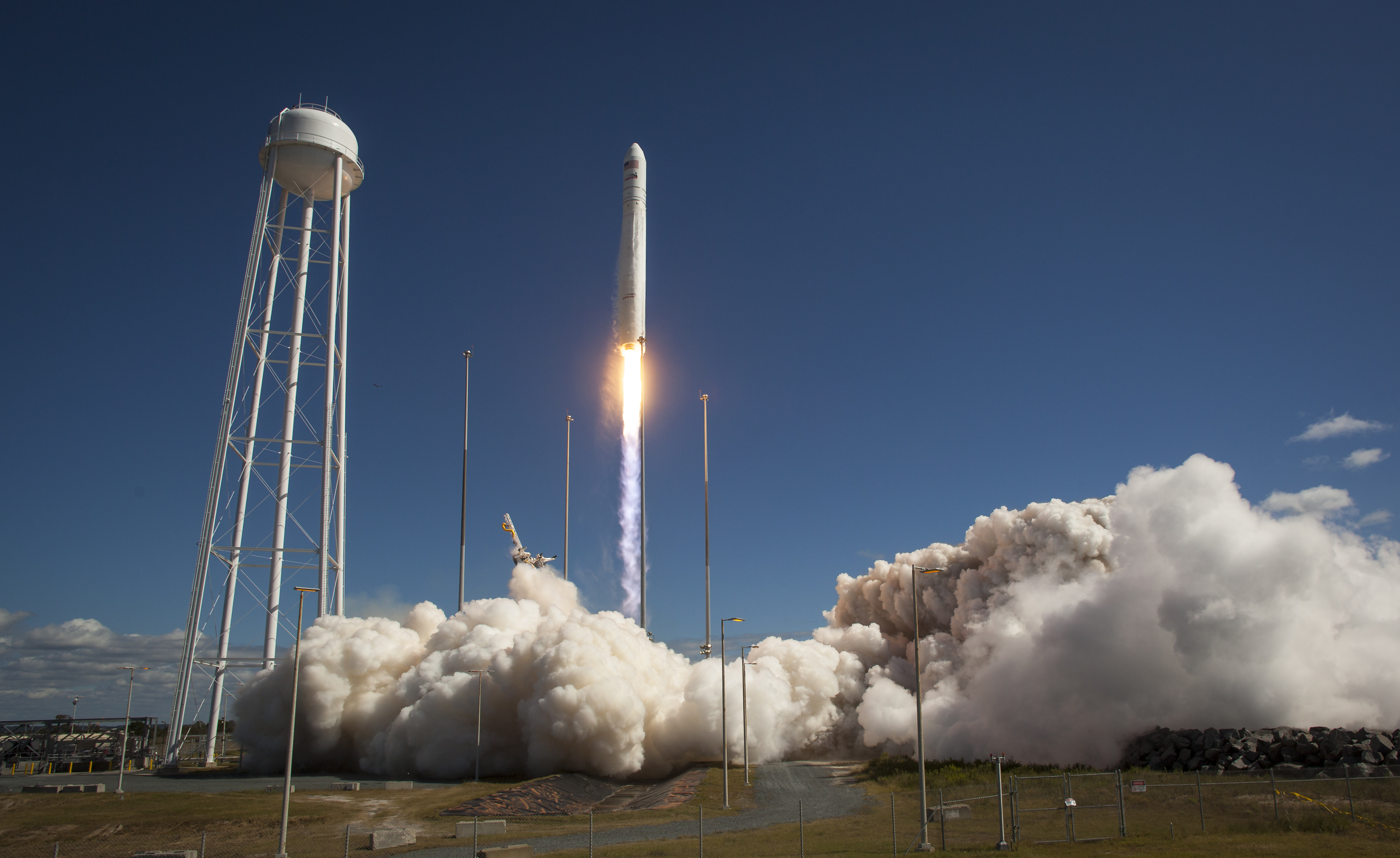 The Antares rocket launching from Pad-0A with a bright white plume underneath against a mostly clear blue sky. A large water tower is stands to the left.