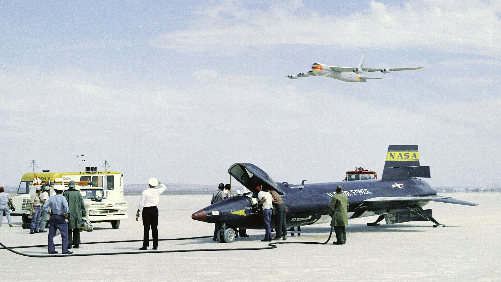 As crew members secure the X-15 rocket-powered aircraft after a research flight, the B-52 mothership flies overhead.