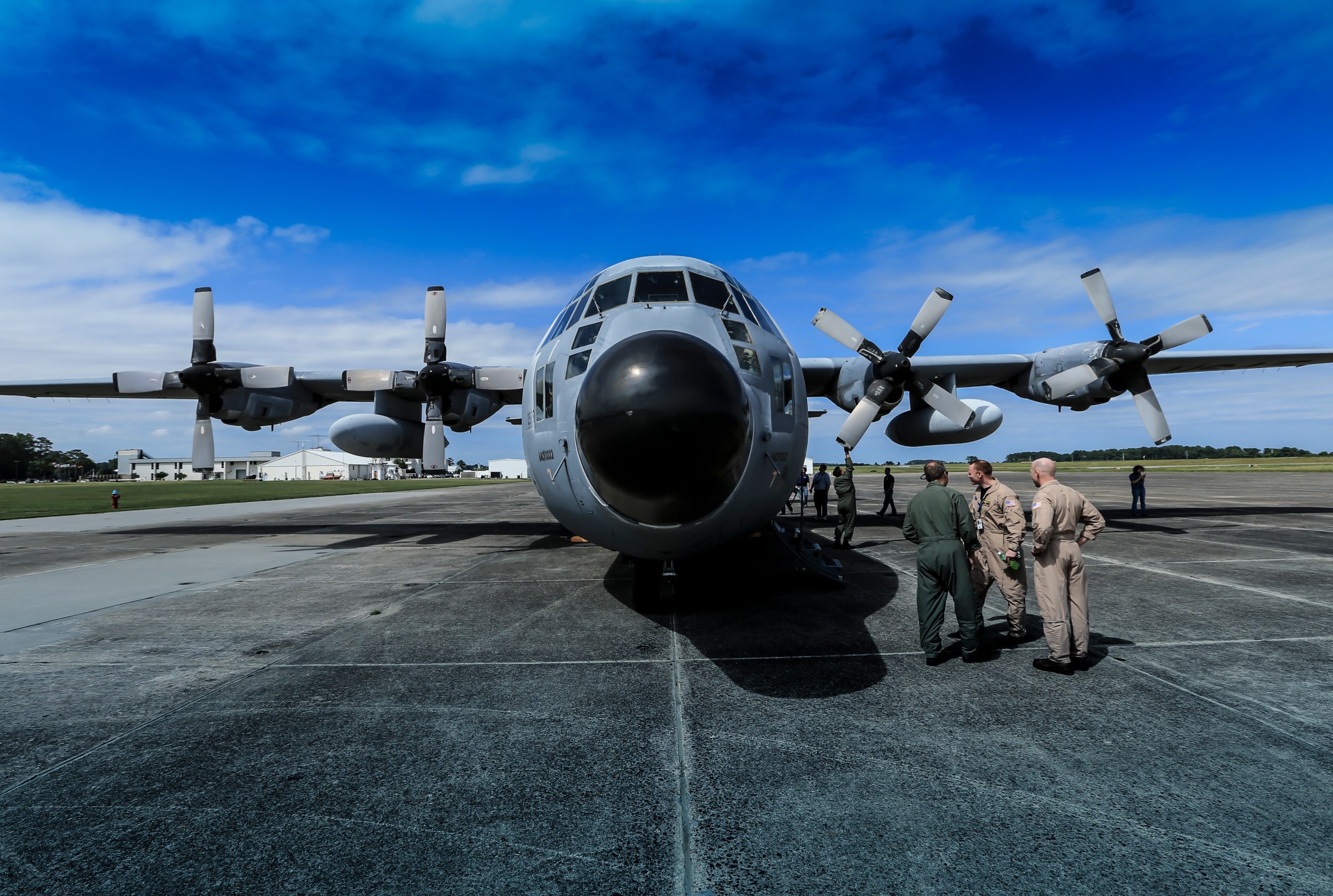 A head-on photo of a C-130 on a runway at Wallops Flioght Facility. the C-130 has four propeller engines on the wings, with a fuel tank mounted on each wing in the center under the wing. the whole plane is a slate, blue-gray color. In the foreground to the right of the plane are two men in khaki jumpsuits talking to a man in a green jumpsuit. Five men are in the shadow of the plane on the right side, interacting with various machines and plane parts.